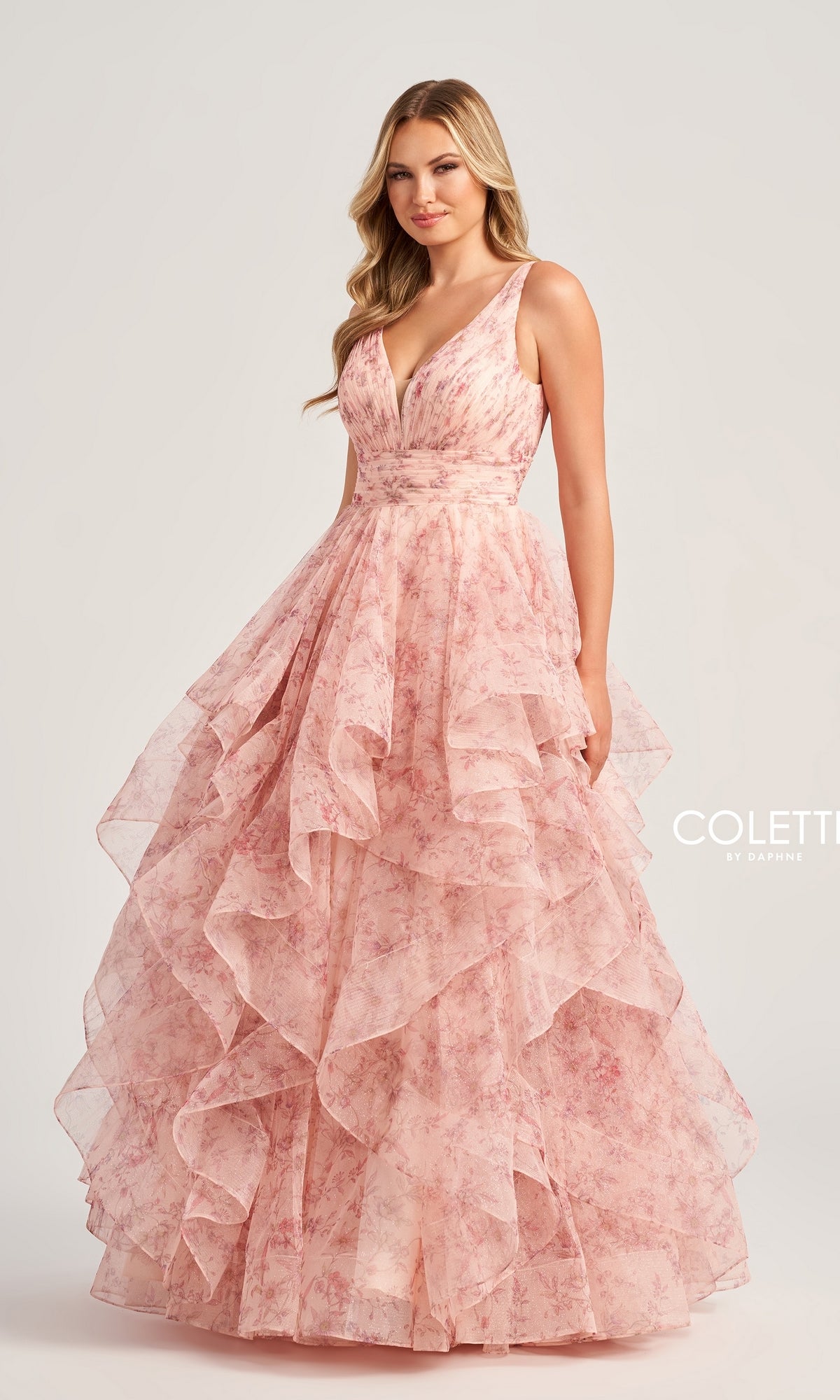 A woman wears the Colette Tiered Long Print Prom Ball Gown CL5273, a sleeveless pink floral dress with a deep V-neckline and layered ruffles, standing against a plain light background.